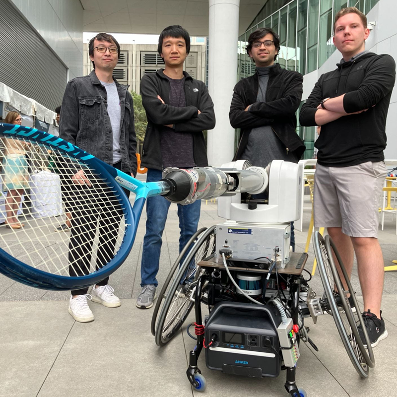 four researchers standing around a robot wheelchair with a tenis racket attached and pointing outward