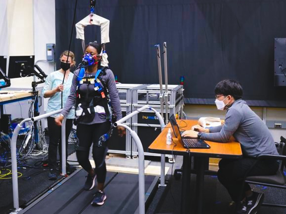 a women with measuring equipment on her body is walking on a treadmill. Two researchers are observing visually and on a computer