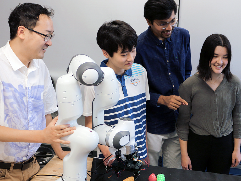 Professor and students standing around a robotic arm looking at a computer